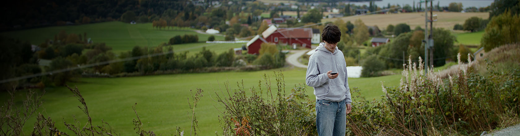 Gutt står og ser på mobilen sin med natur og rural bebyggelse rundt seg