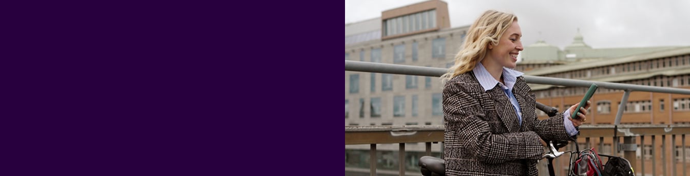 Ung kvinne i kåpe står på en bro i byen med sykkelen sin og ser på mobilen mens hun smiler.