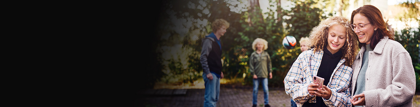 Familie ute i hagen på en solrik høstdag. Mor og datter smiler og ser på en mobiltelefon.