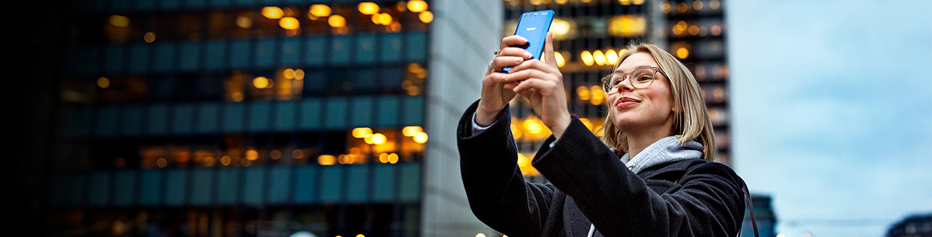 Kåpekledd kvinne utenfor næringsbygg står og tar selfie med mobilen med et smil om munnen