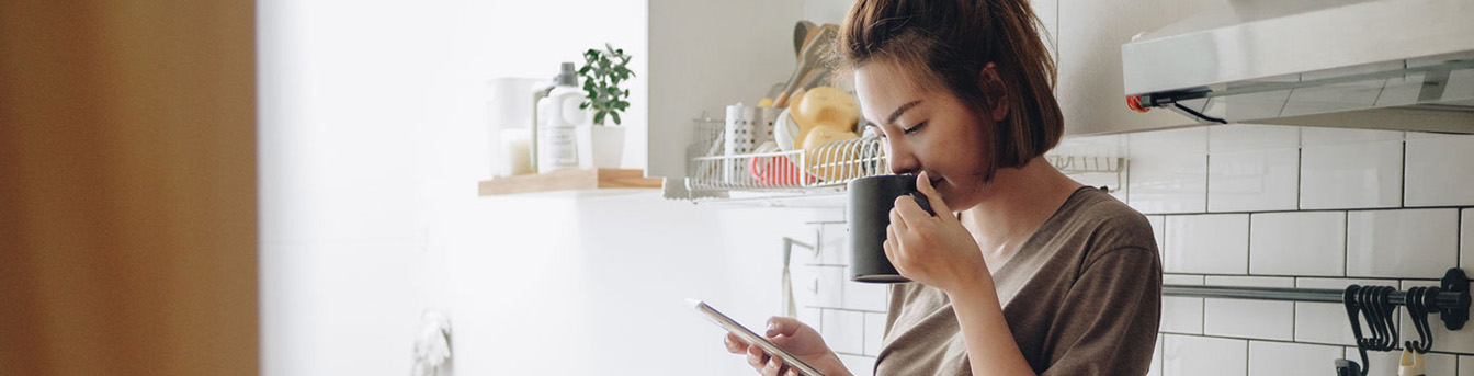 Ung kvinne drikker kaffe og ser på mobilen, sittende på kjøkkenbenken.