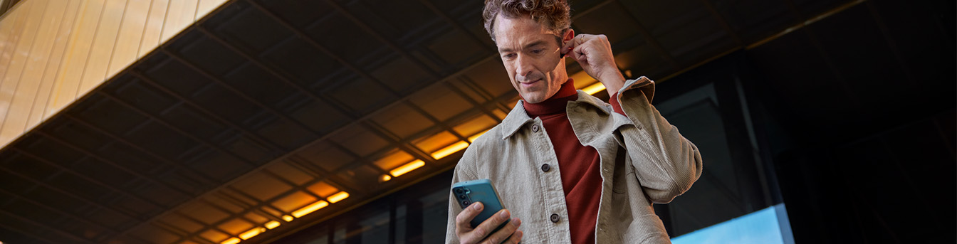 En mann i beige jakke står foran en bygning og ser på mobiltelefonen mens han fester en ørepropp