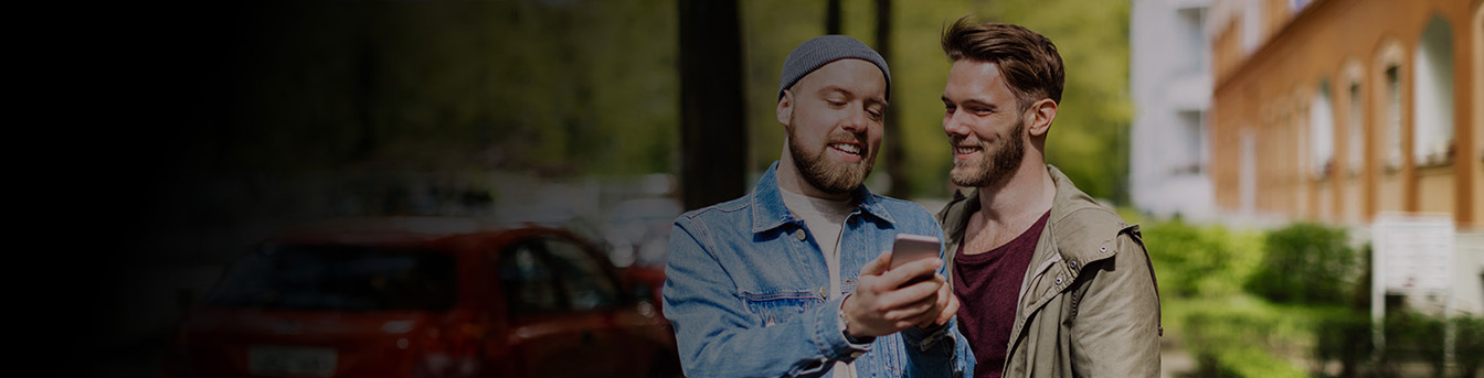 To menn står på gata sammen en fin vårdag, de smiler og ser på en mobil sammen.