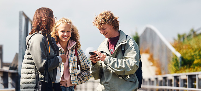 Mor og barn står på en bro og smiler, gutten ser på mobilen.