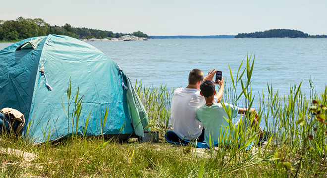 Gutter ved sjøen har satt opp teltet og tar selfie mens sola skinner og speiler seg i vannet