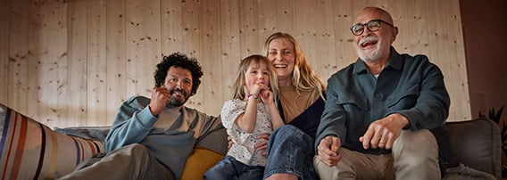 Glad familie sitter og ser TV i sofaen, med popcorn på bordet og smil om munnen.