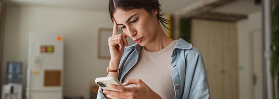 Ung kvinne står i stua og ser på mobilen med et litt usikkert ansiktsuttrykk.